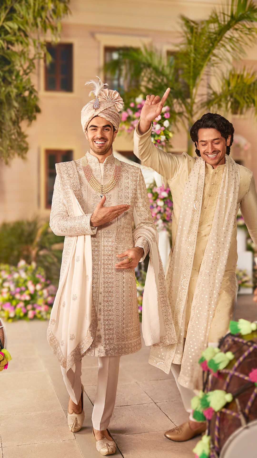 Smiling groom in an intricately embroidered cream sherwani with layered pearl necklaces and turban, walking joyfully with a friend in a beige kurta pajama and embroidered stole